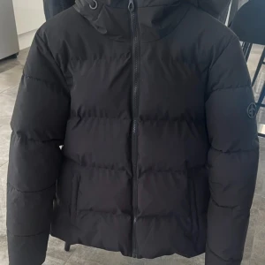Svart pufferjacka från Cross Sportswear - Säljer en svart pufferjacka från Cross Sportswear i storlek L. Jackan har huva, dragkedja framtill och diskret logga på ärmen. Perfekt för kalla dagar med sin vadderade design och stilrena look.