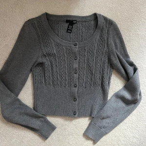 Grå kofta med knappar från H&M - Supersöt grå kofta från H&M. Croppad modell med rund hals, långa ärmar och snygga knappar framtill. Framsidan har ett detaljerat hålmönster som ger en extra touch. Perfekt att slänga över en topp eller klänning.