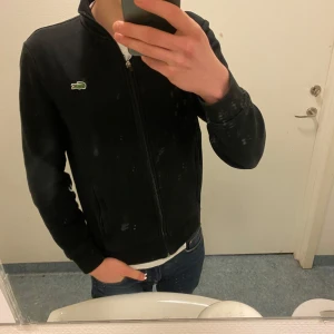 Svart långärmad zip up från Lacoste - Säljer då en svart zip up från Lacoste med dragkedja och det klassiska krokodilmärket på bröstet. Tröjan har en normal passform och är perfekt för en stilren look. Passar både bra till vår eller sommarkvällar! Kom med bud 