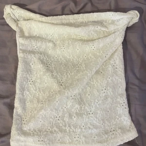 Vit spetsig ärmlös topp med knut - Säljer en vit ärmlös topp i spets med snygga knutdetaljer på axlarna. Toppen har en lätt transparent look och ett romantiskt mönster i tyget. Perfekt för dig som gillar en feminin och trendig stil. Sitter croppad och har en avslappnad passform.