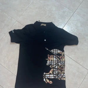 Svart Burberry t-shirt med tryck - Svart t-shirt från Burberry med klassiskt rutigt tryck i beige, svart och rött på framsidan som föreställer en riddare. T-shirten har korta ärmar, krage med knappar och broderad Burberry-logga på ärmen. Materialet är mjuk bomull och passformen är normal.