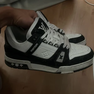Louis Vuitton svartvita sneakers - Snygga Louis Vuitton sneakers i svart och vitt med tydliga LV-loggor på tunga, häl och sula. Skorna har snörning, platt sula och är tillverkade i skinn med präglade detaljer och kontrasterande paneler. Perfekta för dig som vill sticka ut med lyxig streetstyle.