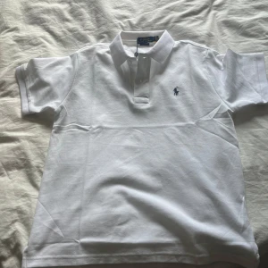 Vit pikétröja från Ralph Lauren - Klassisk vit pikétröja från Polo Ralph Lauren i bomull. Tröjan har korta ärmar, krage och knäppning framtill samt den ikoniska broderade loggan i blått på bröstet. Perfekt för en clean och stilren look.