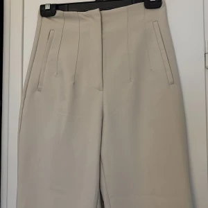 Beige kostymbyxor  - Stilrena beige kostymbyxor med hög midja och raka ben. Byxorna har pressveck framtill och två snedställda fickor. Materialet känns mjukt och har en lätt stretch. Tyvär för små för mig. 