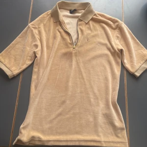 Beige pikétröja i sammet med half zip - beige pikétröja i mjuk sammet med klassisk krage och half zip-dragkedja framtill. Tröjan har korta ärmar och en diskret broderad logga på bröstet. Den har några fläckar här och där, men de syns bara väldigt nära. Står xxs men passar också xs-s