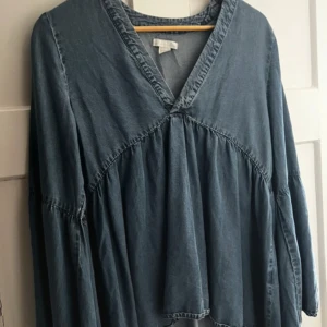 Blå långärmad klänning från H&M - En avslappnad blå blus från H&M med v-ringning och lång ärm. Klänningen har en lös passform och är sydd i mjukt bomullsliknande material. Perfekt för dig som gillar en chill och enkel stil. 