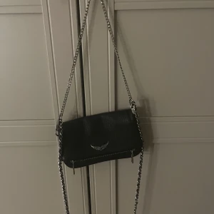 Svart axelväska Zadig & Voltaire skinn - Snygg svart axelväska från Zadig & Voltaire i äkta skinn med silverfärgad kedjerem. Väskan har en ikonisk vingedetalj framtill, dragkedja både fram och bak samt stilren design som passar till allt. Perfekt storlek för det viktigaste. (Den har sminkfläckar men de är i insidan av väskan så de syns ej)