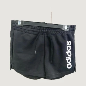 Svarta Adidas shorts med snörning - Svarta sportiga shorts från Adidas med vit logga längs ena benet. De har elastisk midja med snörning och är tillverkade i mjukt material som passar perfekt för träning eller chill. Klassisk och enkel design med korta ben och bekväm passform.