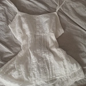 Vit broderad linnetopp med knappar - Superfin vit linnetopp med broderade blommor och knappar framtill. Toppen har smala axelband och är ärmlös, perfekt för varma dagar. Materialet är lätt och luftigt, med fina spetsdetaljer och broderier som ger en romantisk vibe.