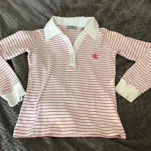 Randig pikétröja från Henri Lloyd - Vit och röd randig pikétröja från Henri Lloyd med klassisk krage och långa ärmar. Tröjan har smala röda ränder, broderad logga på bröstet och breda muddar vid ärmsluten. Perfekt för en sportig och preppy look.