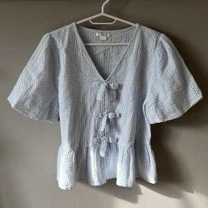 Ljusblå blus med knyt från Gina Tricot - Supersöt ljusblå blus från Gina Tricot med v-ringning och tre knyt framtill. Blusen har korta ärmar, volang nertill och ett diskret randigt mönster i strukturen. Perfekt för en fräsch och somrig look! Den är från Gina Young! Använd endast 1 gång.
