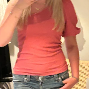 Korallrosa blus med puffärm - Säljer en korallrosa blus med korta puffärmar och rundad hals. Toppen har en lätt rynkad struktur i tyget som ger en cool känsla och sitter figurnära. Perfekt att styla med jeans för en fräsch look.