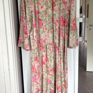 Blommig långklänning från Lindex - Långklänning från Lindex med beige botten och färgglatt blommigt mönster i rosa, grönt och orange. Klänningen har långa ärmar och avslappnad passform, perfekt för sommarens alla tillfällen. Tillverkad i mjuk bomull som känns skön mot huden.
