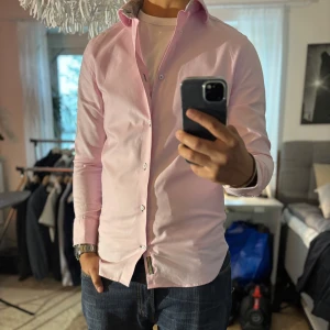 Ljusrosa skjorta från Riley, slim fit - Säljer en ljusrosa skjorta från Riley i slim fit-modell. Skjortan har långa ärmar, vita knappar och snygga detaljer med blommigt mönster på insidan av kragen och manschetterna. Perfekt för dig som gillar stilrena plagg med en twist.