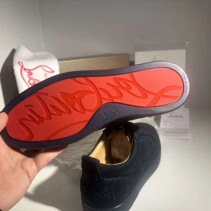  Christian Louboutin Skor - Exklusiva mörkblå sneakers från Christian Louboutin i mocka med ikonisk röd sula och ton-i-ton broderad logga på hälen. Skorna har rund tå, platt sula och klassisk snörning. Insidan är ljusbeige och materialet känns lyxigt och mjukt.