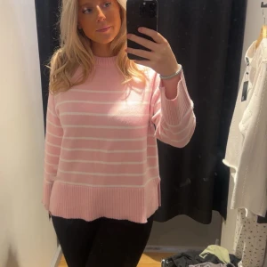 Rosa randig stickad tröja - Superfin rosa stickad tröja med vita ränder och bred ribbad kant nedtill. Tröjan har en avslappnad passform och långa ärmar, perfekt för att mysa i under kyligare dagar. Snygg till både jeans och kjol!
