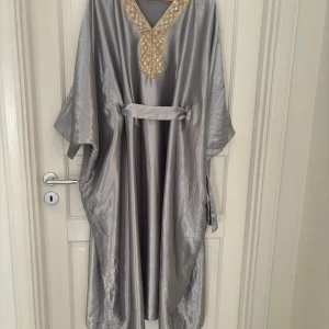 Silvrig långklänning från Shein - Säljer en glansig silvrig långklänning från Shein med v-ringning och vackra guldfärgade paljettdetaljer vid halsen. Klänningen har lös passform, bälte i midjan och vida ärmar. Perfekt för dig som vill sticka ut med en elegant look.