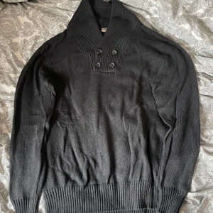 Svart stickad tröja med knappar - Svart stickad tröja från Livergy med bred ribbad krage och fyra dekorativa knappar framtill. Tröjan har lång ärm och bred ribbad mudd vid ärmslut och nederkant. Perfekt för dig som gillar en stilren och enkel look.