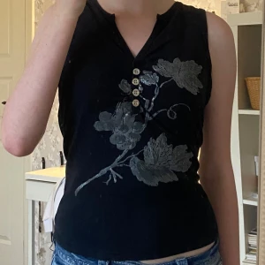 Svart blommig linnetopp från b.young - Svart ärmlös linnetopp från b.young med snyggt silvergrått blommönster framtill. Toppen har v-ringning med dekorativa knappar och en snygg passform. Perfekt för dig som gillar toppar med snygga detaljer, eller vill ha något unikt i garderoben 💋😍