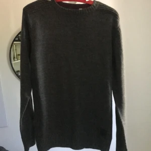 Mörkgrå stickad tröja - En enkel och stilren mörkgrå stickad tröja med rund halsringning och långa ärmar. Tröjan har ribbade muddar vid ärmslut och nederkant, vilket ger en snygg och klassisk look. Perfekt att matcha med jeans eller chinos.