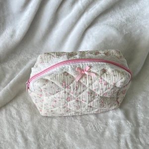 Quiltad blommig necessär med rosett - Supergullig quiltad necessär i ljusbeige bomullstyg med små rosa blommor, spetsdetaljer och en söt rosa rosett framtill. Dragkedjan är rosa och insidan är fodrad i matchande rosa. Perfekt för smink eller småprylar!