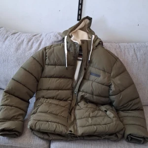 Olivgrön pufferjacka från Blend XL - Säljer en olivgrön pufferjacka från Blend i storlek XL. Jackan har huva med vita snören, mysigt teddyfoder på insidan och dragkedja med tryckknappar. Perfekt för kalla dagar när du vill hålla värmen och ändå se chill ut.
