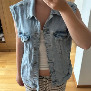 Ljusblå jeansväst med knappar - Säljer en snygg ljusblå jeansväst med klassisk krage och knäppning framtill. Västen har två bröstfickor med lock och knappar, samt en rak och lite loose passform. Perfekt att slänga över en t-shirt för en chill look. Köpt secondhand från marker garcia