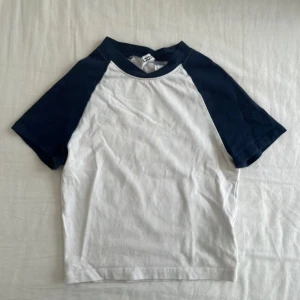 Vit och marinblå top - Vit top med marinblå raglanärmar från H&M. Klassisk basebollstil med rund halsringning och kontrastfärgade ärmar. Perfekt för en chill och sportig look. Gjord i mjuk bomull som känns skön mot huden.