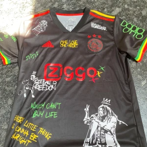 Ajax x Bob Marley fotbollströja Adidas - Unik svart Ajax fotbollströja från Adidas med Bob Marley-tema. Färgglada detaljer i rött, gult och grönt på ärmarna, tryck med texter och illustrationer, samt Ajax-logga och Ziggo sponsor. Materialet är lätt och andas, perfekt för fotboll eller streetwear.