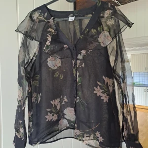 Blommig Visual Clothing Project blus  - Snygg svart transparent skjorta från Visual Clothing Project med blommigt mönster i ljusa toner. Skjortan har v-ringning, knappar framtill och långa ärmar. Perfekt för dig som vill sticka ut med en trendig och luftig look. Underbar Visula Clothing project blus i skirt tyg på armar men täckande över kroppen. Volang krage och så fin 😍 är M och passar S också. Finare i verkligheten ☺️