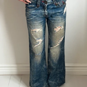 Blåa bootcut jeans med slitningar - Supersnygga jeans med låg midja och utsvängda ben. Modellen har en figurnära passform upptill och en lätt bootcut. Jeansen är i ett tvättat blått denimtyg med kraftiga slitningar över lår och knän. Det saknas en knapp på bakfickan (sista bilden), detta är dock inget som man lägger märke till.  Mått: Innerben – 67 cm Ytterben – 90 cm Midjemått rakt över – 35 cm Midjehöjd – 21 cm Benvidd nertill – 25 cm  Jag är 161 cm. Detta är en återpublicerad annons från en annan sida, priset är diskuterbart.