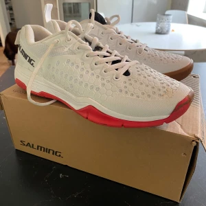 Vita Salming sneakers med röd sula storlek 41 1/3 - Använda bara en gång. Snygga vita sneakers från Salming med hexagonmönstrad ovandel och cool röd och brun sula. Skorna har snörning och detaljer i svart samt en liten svensk flagga på plösen. Perfekta för träning eller sportiga outfits.