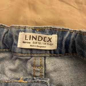 Ljusblå jeansshorts från Lindex - Snygga ljusblå jeansshorts från Lindex med klassisk femficksdesign, uppvikta benslut och bekväm resår i midjan. Shortsen har bälteshällor och knappstängning framtill. Perfekta för varma dagar och enkel att matcha med allt.
