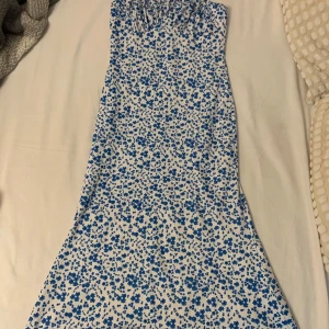 Blommig långklänning i blått och vitt - Supersöt långklänning med blått blommönster på vit botten. Klänningen är ärmlös och har en smockad överdel som sitter snyggt. Perfekt för sommarens alla tillfällen och ger en fräsch och trendig look.