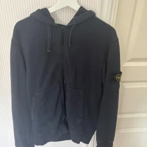 Mörkblå zip hoddie från Stone Island - Mörkblå hoodie från Stone Island med dragkedja framtill och klassisk patch på ärmen. Tröjan har huva med snörning och två stora fickor framtill. Tillverkad i mjuk bomull, perfekt för en chill och stilren look.