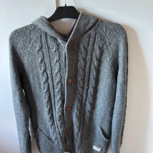 Populär Grå stickad cardigan med huva - Säljer en grå stickad kofta med huva och snygga träknappar. Koftan har kabelstickat mönster framtill, två fickor och långa ärmar. Perfekt för lager-på-lager och chill dagar. Passar dig som gillar en avslappnad och mysig stil. Perfekt för sena sommarkvällar! 