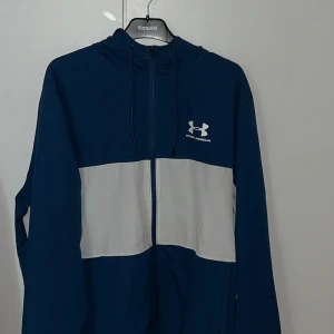 Blå och vit vindjacka från Under Armour - Snygg blå och vit vindjacka från Under Armour med dragkedja och huva. Riktigt sällsynt jacka. Strl XL men passar även L. Jackan är oanvänd och helt felfri. Hör av er vid frågor!