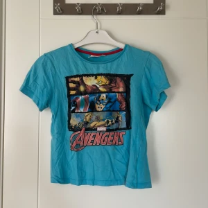 Blå Marvel Avengers t-shirt - Cool blå t-shirt med tryck av Avengers-hjältarna Iron Man, Captain America och Thor på framsidan. T-shirten har rund hals med röd kant och är kortärmad. Perfekt för dig som älskar superhjältar och vill sticka ut med en färgstark look. Tröjan är barnstorlek men passar som XS/S. 