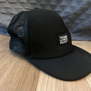 Svart keps  - Svart keps från The Ultimate Project med nät baksida och justerbar storlek. Framsidan har en svart patch med vit text. Kepsen är tillverkad i 100% polyester. 