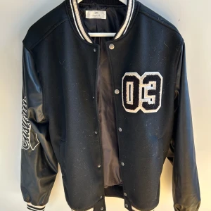 Svart bomberjacka från H&M med brodyr - Svart bomberjacka från H&M med stora broderade siffror '03' på bröstet och texten 'Custom' på ena ärmen. Använt 1 gång