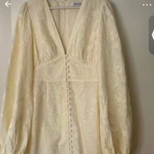 Ljus kortklänning från Adoore - Lånade bilder! Superfin ljus kortklänning från Adoore med djup v-ringning, puffiga långärmar och dekorativa knappar framtill. Klänningen har ett subtilt blommigt mönster och är sydd i ett lätt, elegant tyg som ger en lyxig känsla.