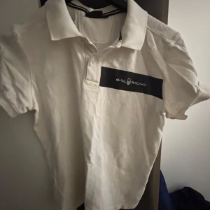 Vit pikétröja från Sail Racing - Vit pikétröja från Sail Racing med klassisk krage och korta ärmar. Tröjan har svart logotypdetalj på bröstet och en liten broderad detalj i nacken. Tillverkad i bomull, perfekt för en clean och sportig stil.