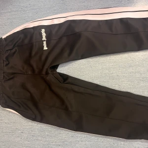 Palm Angels svarta mjukisbyxor - Svarta mjukisbyxor från Palm Angels med vita sidoränder och broderad logga på låret. Byxorna har elastisk midja och är tillverkade i ett glansigt syntetmaterial som ger en sportig vibe. Perfekta för en avslappnad streetwear-look.