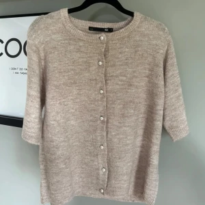 Beige stickad kofta med pärlknappar - Supermjuk beige kofta från 157 med ribbad rund halsringning och pärlknappar framtill. Koftan har halvkort ärm och är stickad i ett fluffigt material som känns både chill och stilrent. Perfekt att slänga över en t-shirt eller bära som den är.
