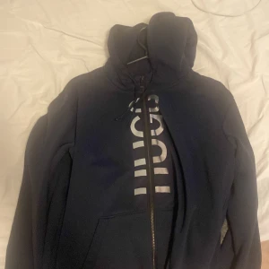 Mörkblå hoodie från Hugo Boss - Säljer en mörkblå hoodie från Hugo Boss med vit logga framtill. Tröjan har dragkedja, huva med snörning och känguruficka. Perfekt för dig som gillar stilren streetwear med premiumkänsla.