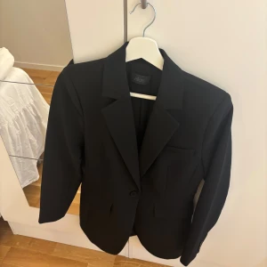 Svart kavaj från Ellos - Klassisk svart kavaj från Ellos med snygg passform, enkelknäppning och tre fickor fram. Kavajen har fyra dekorativa knappar vid ärmslut och är tillverkad i ett slätt material som ger en stilren look. Perfekt för dig som gillar en tidlös och clean stil.