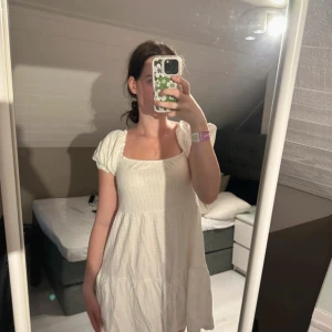 Vit kort klänning med puffärm - Supersöt vit kort klänning med puffärmar och fyrkantig ringning. Klänningen har en flowy passform och är gjord i ett lätt, strukturerat tyg som känns perfekt för varma dagar. Passar dig som gillar en romantisk och avslappnad stil.