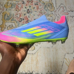 Adidas blå och neon fotbollsskor - Snygga fotbollsskor från Adidas i blått med neonfärgade detaljer i gult och rosa. Skorna har en strömlinjeformad design, låg profil och plös utan snörning. Yttermaterialet är syntet och sulan är grön med dobbar för bra grepp på planen.