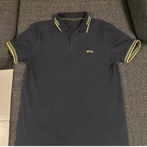 Boss piké  - Snygg mörkblå pikétröja från BOSS med gula ränder på krage och ärmslut. Klassisk polo med korta ärmar, knappar framtill och BOSS-logga på bröstet. Stretchigt material för en bekväm slim fit. Perfekt för en stilren och sportig look.