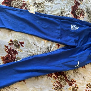 Blå träningsbyxor Puma Marseille - Snygga blå träningsbyxor från Puma med Marseille-logga på ena benet och vita detaljer baktill. Byxorna har smal passform och är perfekta för fotboll eller träning. Tillverkade i mjukt och stretchigt syntetmaterial för maximal rörelsefrihet.